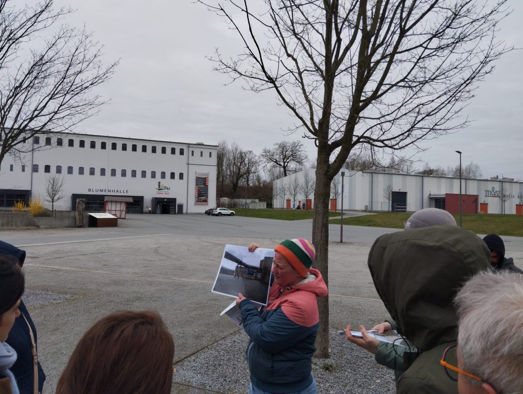 Sprievodkyňa so starými fotografiami vysvetľuje históriu a premeny areálu Landesgartenschau v Löbau pred budovou Blumenhalle, počas študijnej návštevy projektu URBACT GreenPlace.