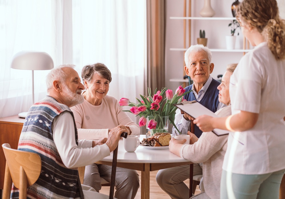 traja seniori sedia za stolom a nad nimi stojí a diskutuje sociálna pracovníčka