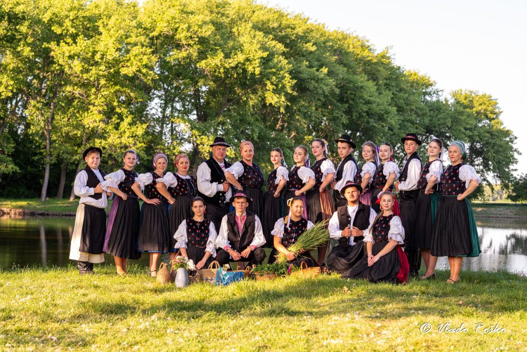 Skupina členov folklórneho súboru v tradičných krojoch pózuje pri jazere na letnej lúke, obklopená košíkmi s bylinkami