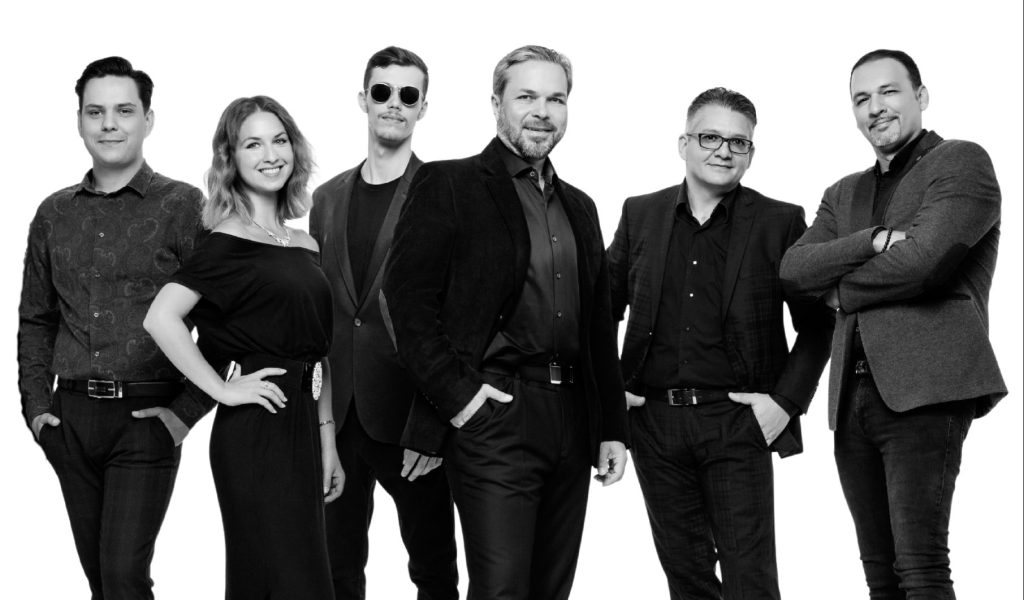 Skupinová fotografia šiestich hudobníkov, ktorí stoja vedľa seba v elegantnom oblečení a uvoľnených pózach na bielom pozadí.