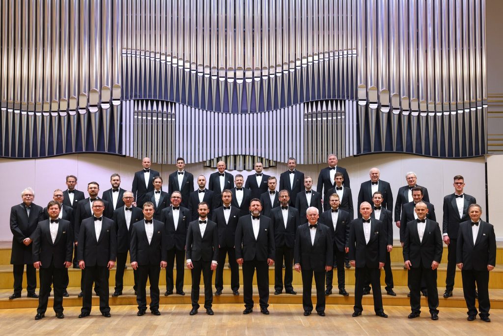 Skupinová fotografia mužského speváckeho zboru v čiernych frakoch stojaceho na pódiu pred veľkolepou stenou organových píšťal.