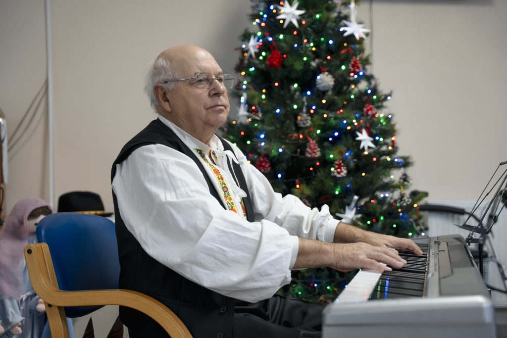 Hudobník hrá na klávesoch pri vianočnom stromčeku v spoločenskej miestnosti zariadenia pre seniorov.