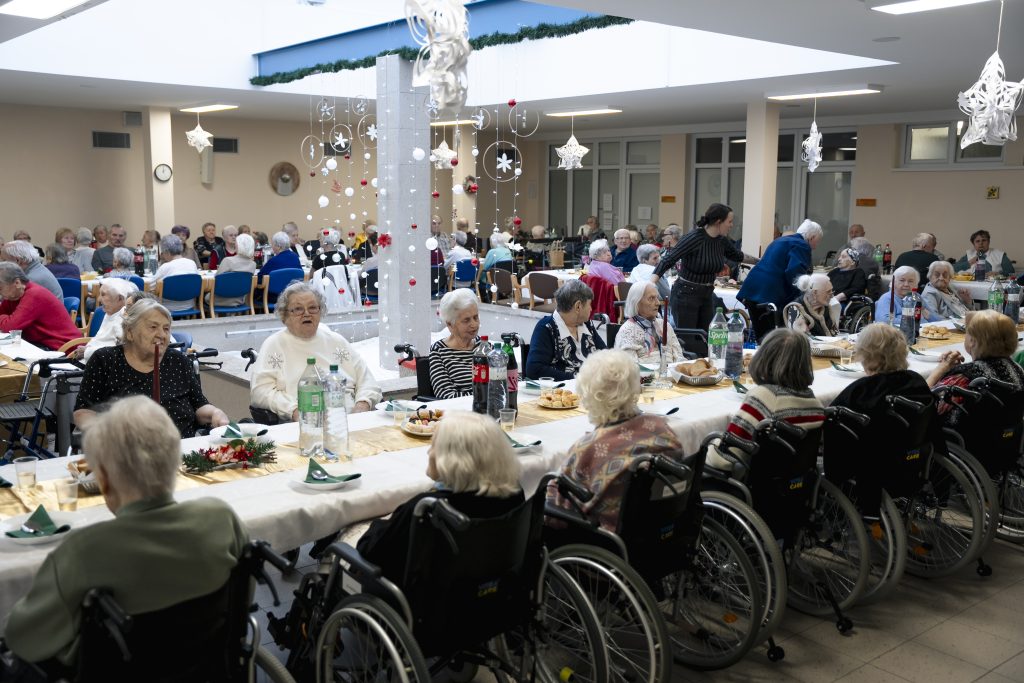 Seniori sedia pri dlhých slávnostne prestretých stoloch počas vianočného posedenia v zariadení sociálnych služieb.