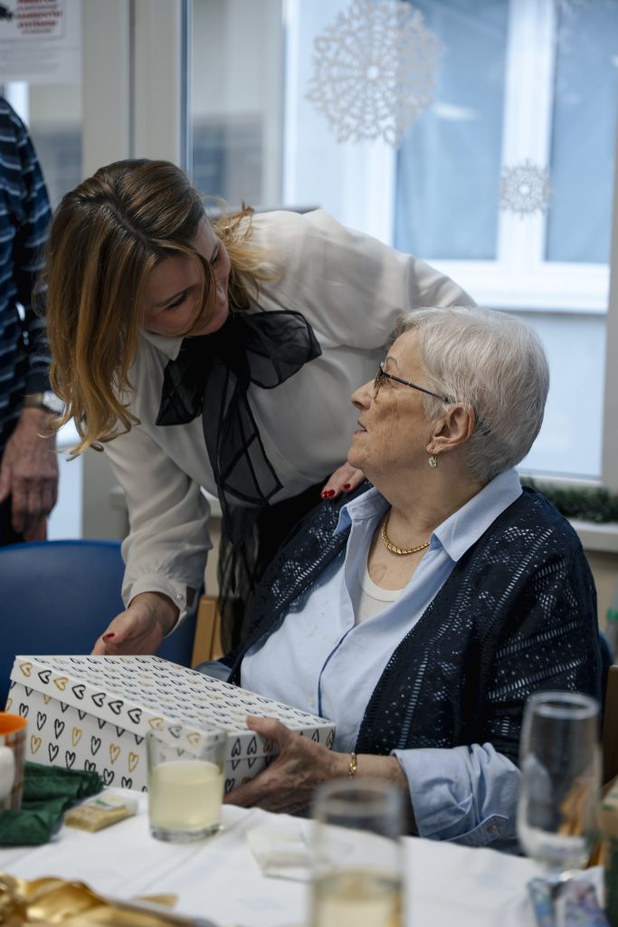 Zamestnankyňa mesta sa rozpráva so seniorkou pri stole a odovzdáva jej darček.