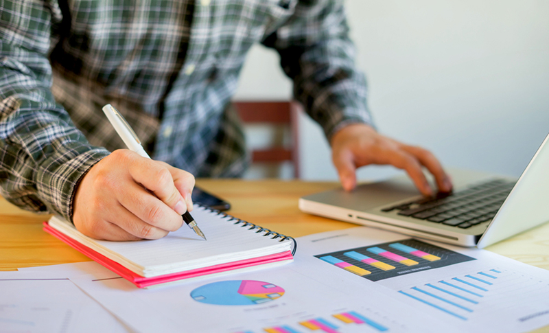 ilustračný záber na mladého podnikateľa, vidno iba jeho vrchnú časť tela, hlavu nie, muž si robí poznámky do notesa, pred sebou má otvorený notebook