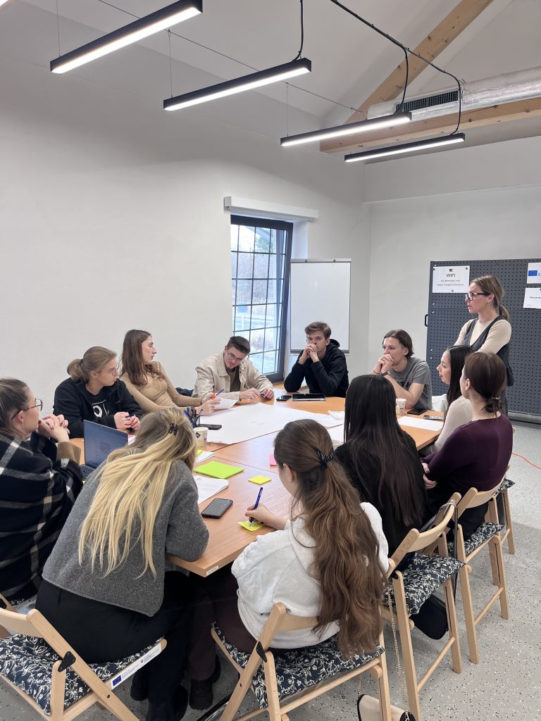 mladí ľudia okolo stola a vyučujúca pri nich stojí