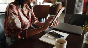 žena drží tablet, na stole má kávu a papiere